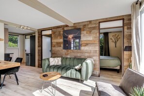 A modern living room with a wooden wall, a sofa, a dining table, and a bed. - Bungalow with large private garden and BBQ place near Lake Veere (Kortgene)
