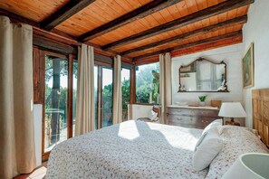 Ein Schlafzimmer mit Holzboden, einem großen Bett mit floralem Bettzeug, einem Schminktisch, einem Spiegel und Blick ins Grüne.