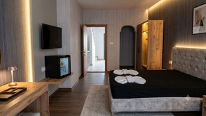 Une chambre d’hôtel moderne dotée d’un téléviseur à écran plat, d’une table de chevet en bois, d’un lit avec des serviettes et d’une grande porte en arc de cercle.