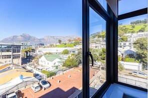 Blick aus einem Fenster auf ein Wohngebiet mit Gebäuden, einen grünen Hügel und Berge in der Ferne.