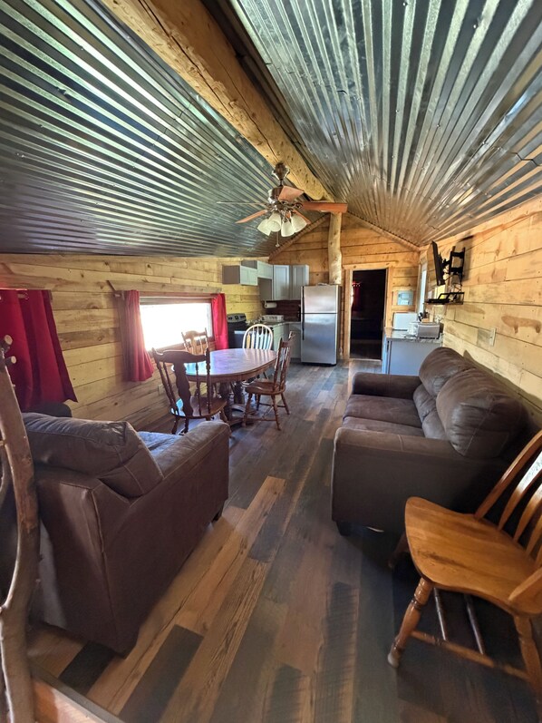 A cabin interior with a dining table, chairs, a sofa, a ceiling fan, and a kitchen area.