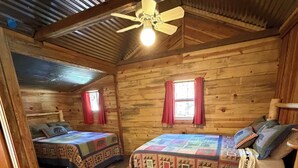 En hytte med to senge i træ, en ventilator i loftet og et vindue med røde gardiner.