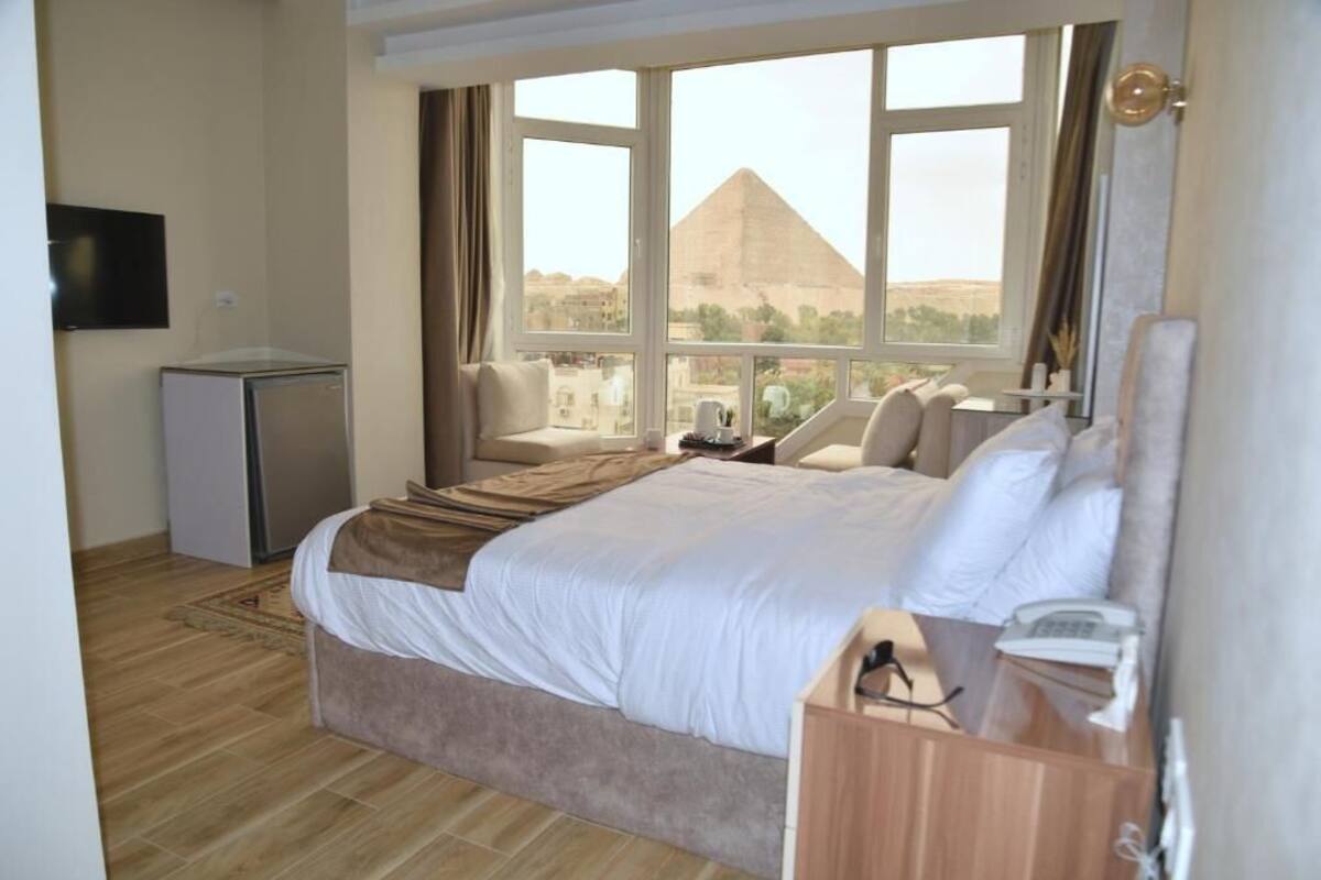 A hotel room with a bed, a desk, a chair, a TV, and a view of a pyramid.
