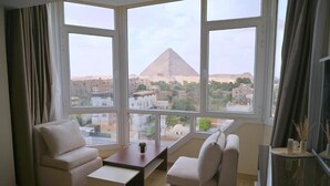 A modern living room with a large window offering a view of a pyramid and surrounding buildings. - Pyramids Wonders Hotel (Giza)
