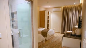A hotel room with a glass shower enclosure, a bed with a brown blanket, a bedside table with a coffee maker, and a wardrobe. - Pyramids Wonders Hotel (Giza)