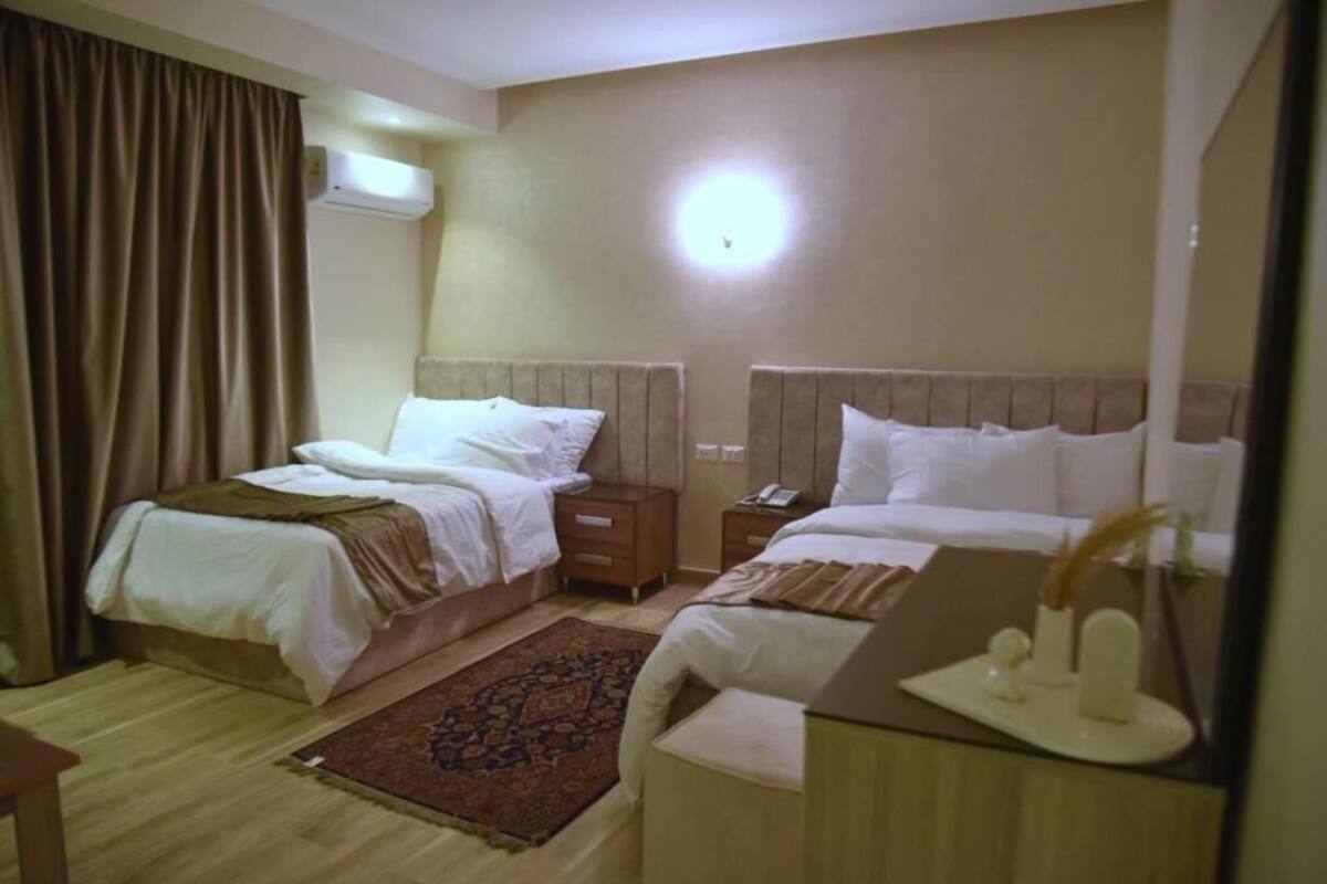 A hotel room with two beds, a bedside table with a lamp, a nightstand, a rug, and an air conditioning unit.