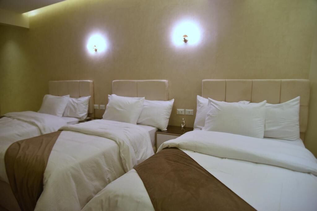 Een hotelkamer met drie bedden, elk voorzien van witte beddengoed en hoofdeinden. Boven de bedden hangen wandlampen.