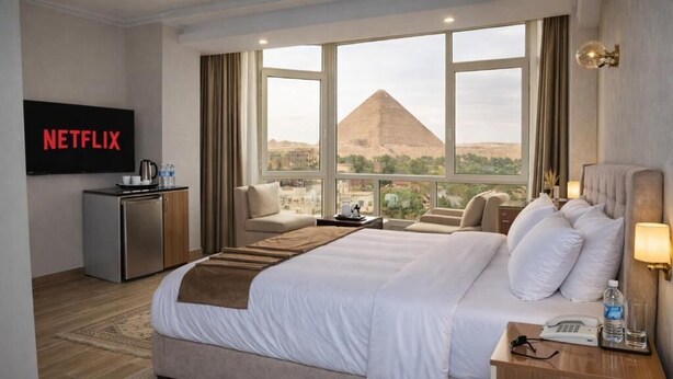 A hotel room with a bed, a TV, a desk, and a view of a pyramid. - Pyramids Wonders Hotel (Giza)