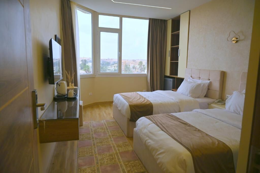 A hotel room with two beds, a TV, a desk with a lamp, and a view of the city.