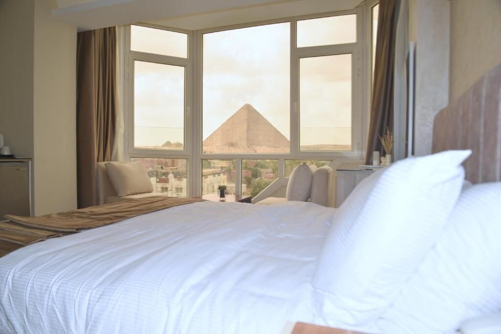 Quarto de hotel com uma cama grande, vista da pirâmide e janela com cortinas.