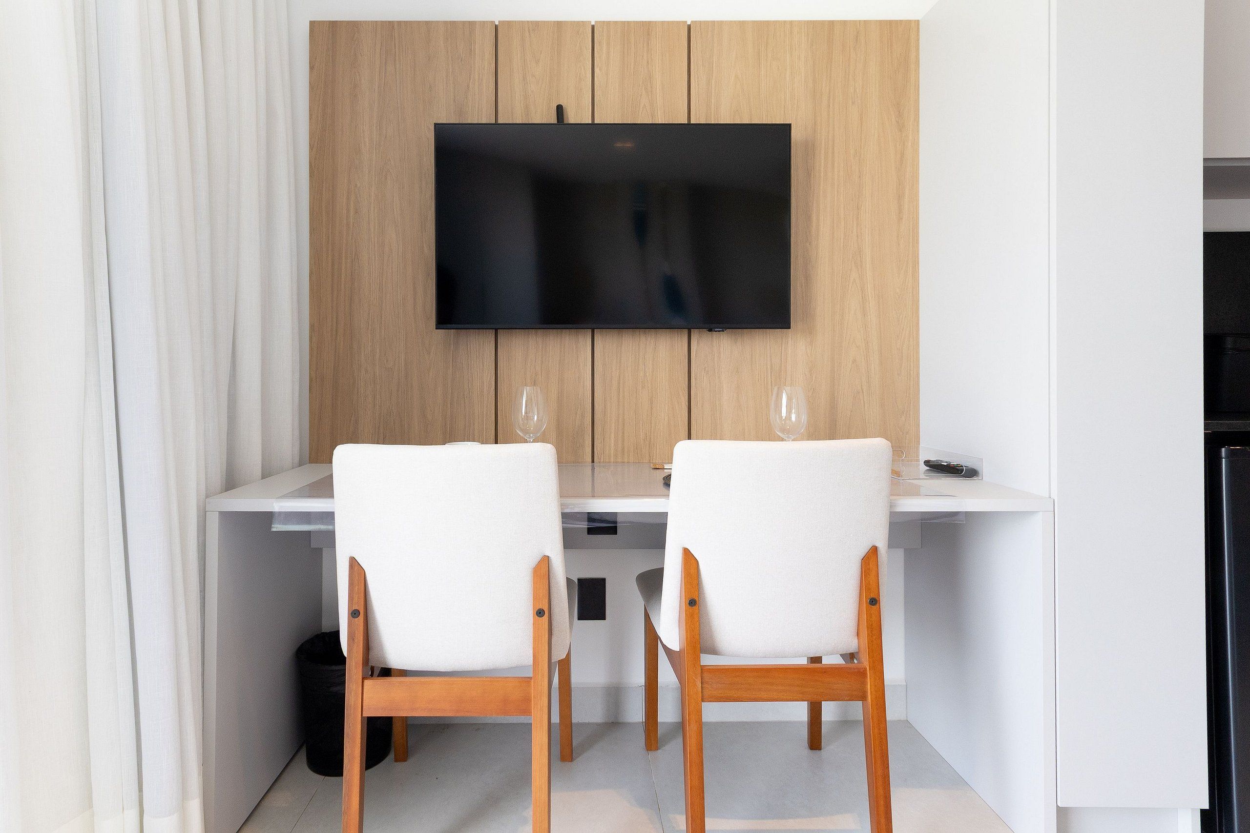 Uma área de refeições moderna com uma televisão de tela plana presa a uma parede de madeira, duas cadeiras brancas e uma mesa branca.