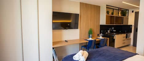 A modern hotel room with a bed, a desk, a TV, a kitchenette, and a chair.