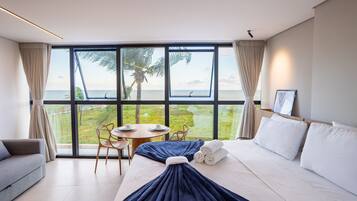 A modern hotel room with a large window, a bed, a sofa, a table with chairs, and a view of the sea.
