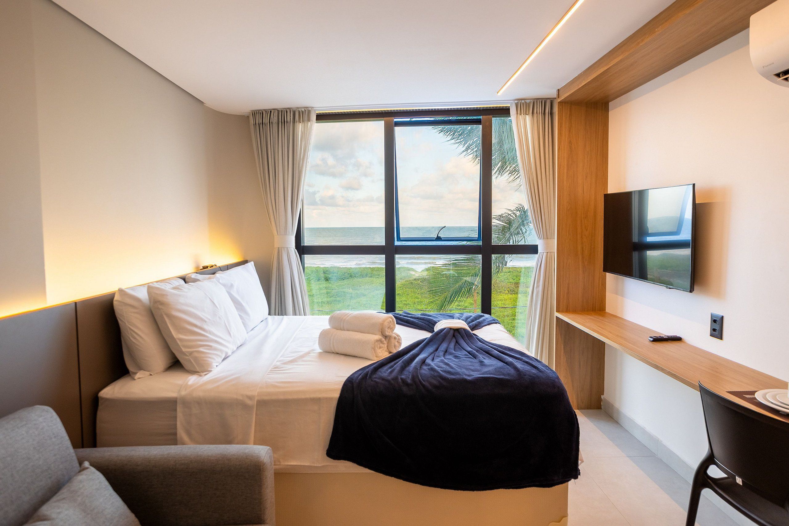Habitación de hotel con cama, escritorio, silla, televisor y un amplio ventanal con vistas al exterior.