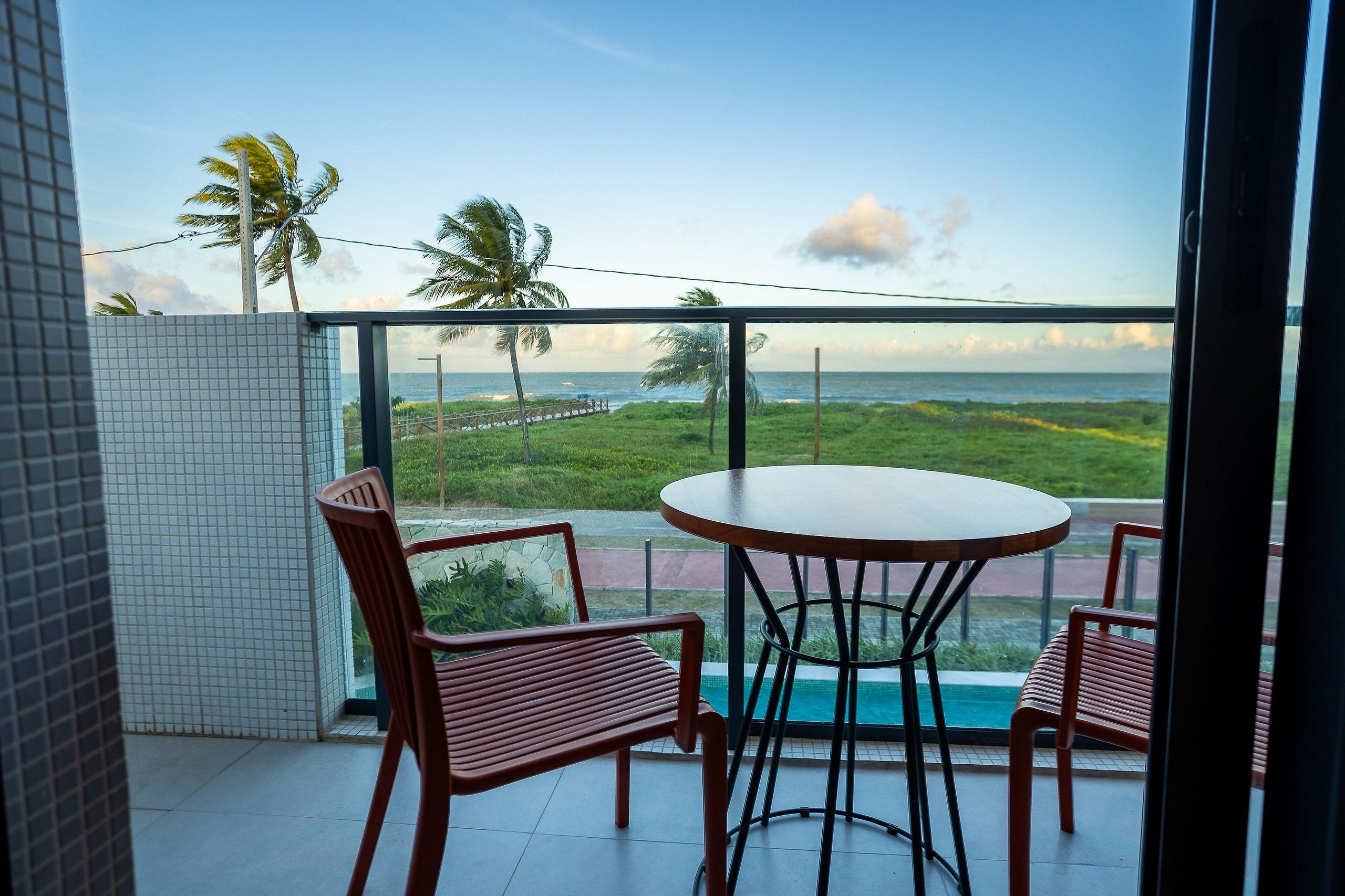 Un balcón con una mesa redonda y dos sillas, con vista a un paisaje costero con palmeras y el mar.