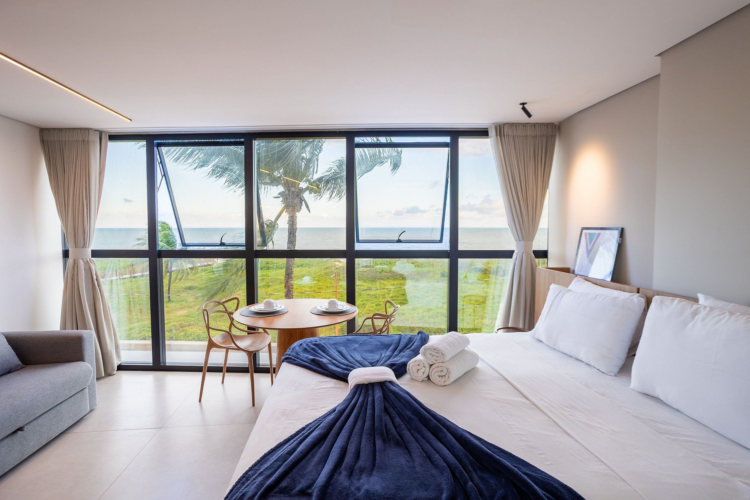 A modern hotel room with a large window, a bed, a sofa, a table with chairs, and a view of the sea.