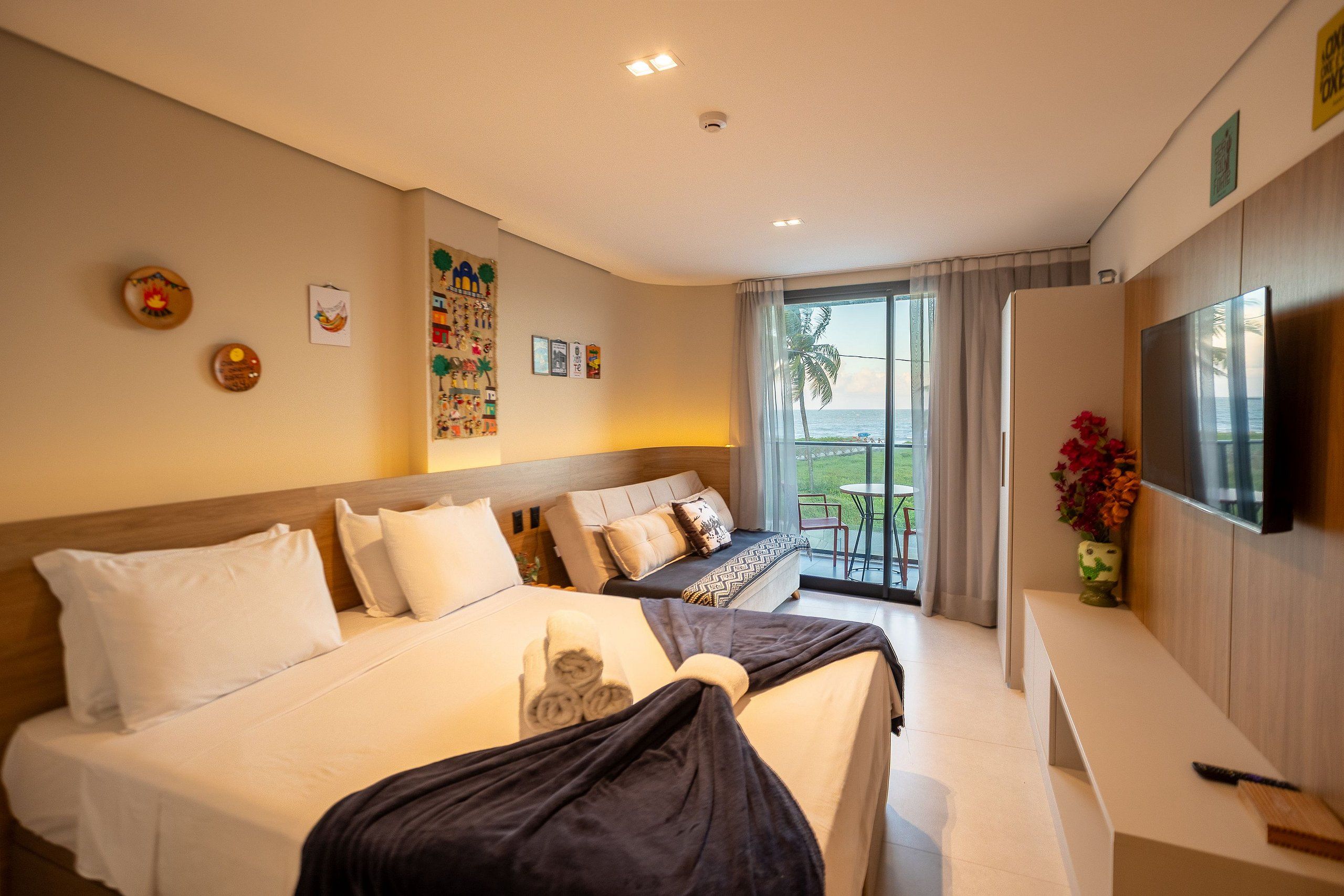 A hotel room with a large bed, a desk, a television, and a balcony with a view of the sea.