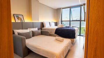 Quarto de hotel moderno com uma cama grande, um sofá e vista para o mar através de amplas janelas.