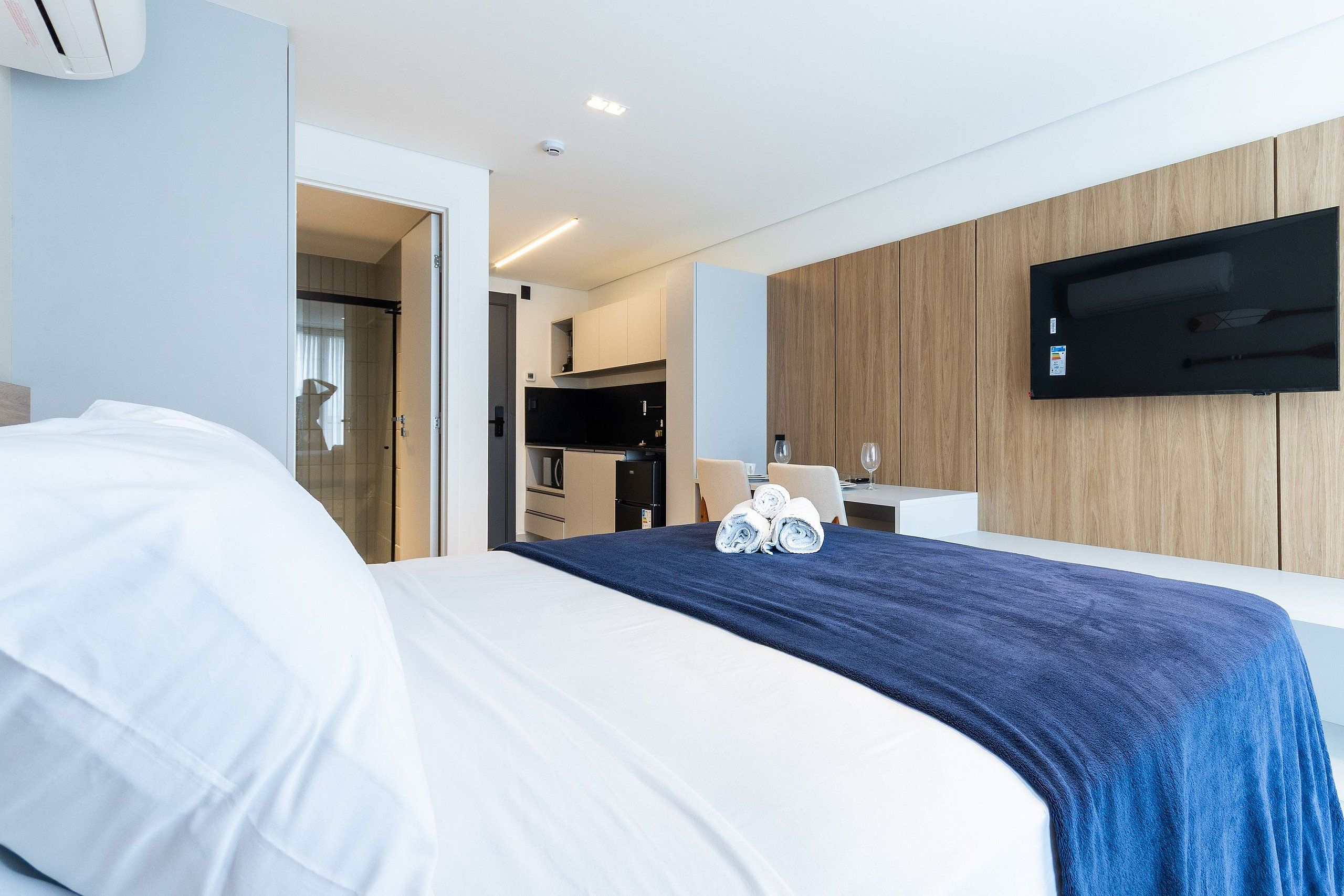 Quarto de hotel com cama, televisão fixada na parede e uma cozinha compacta ao fundo.