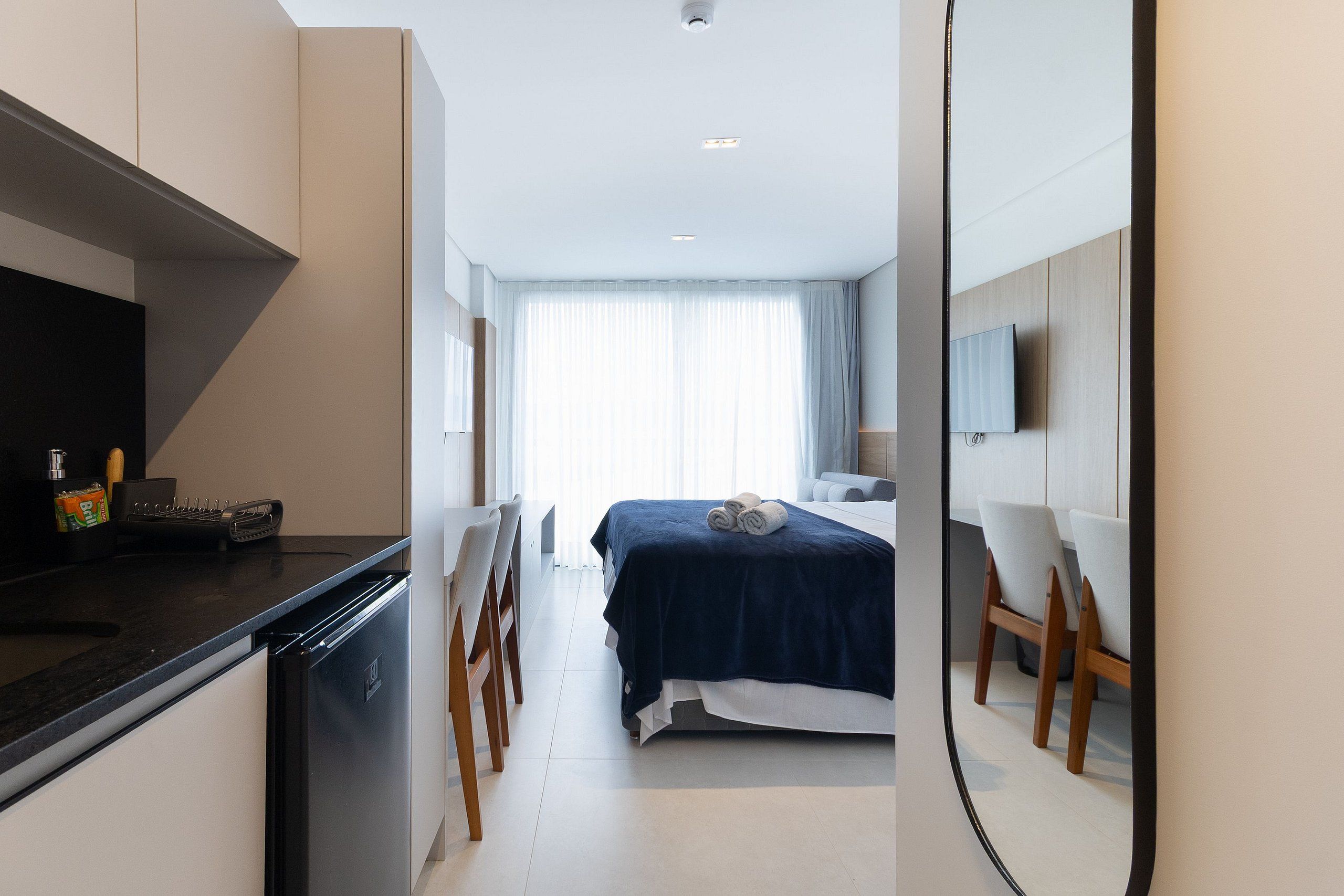 A modern hotel room with a bed, a desk with a computer, a chair, and a small kitchen area.