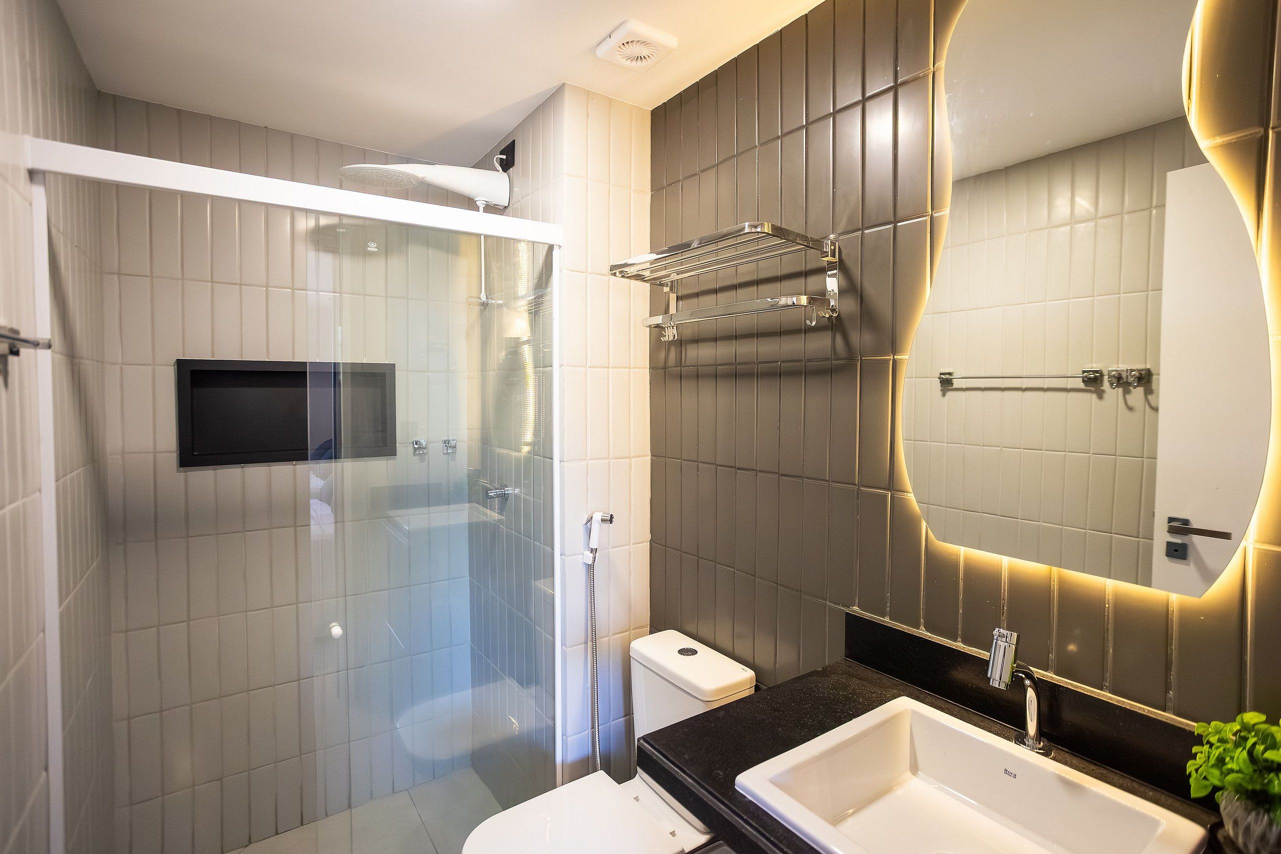 Un baño moderno con ducha envuelta en vidrio, inodoro blanco, encimera negra y un espejo grande con marco iluminado.
