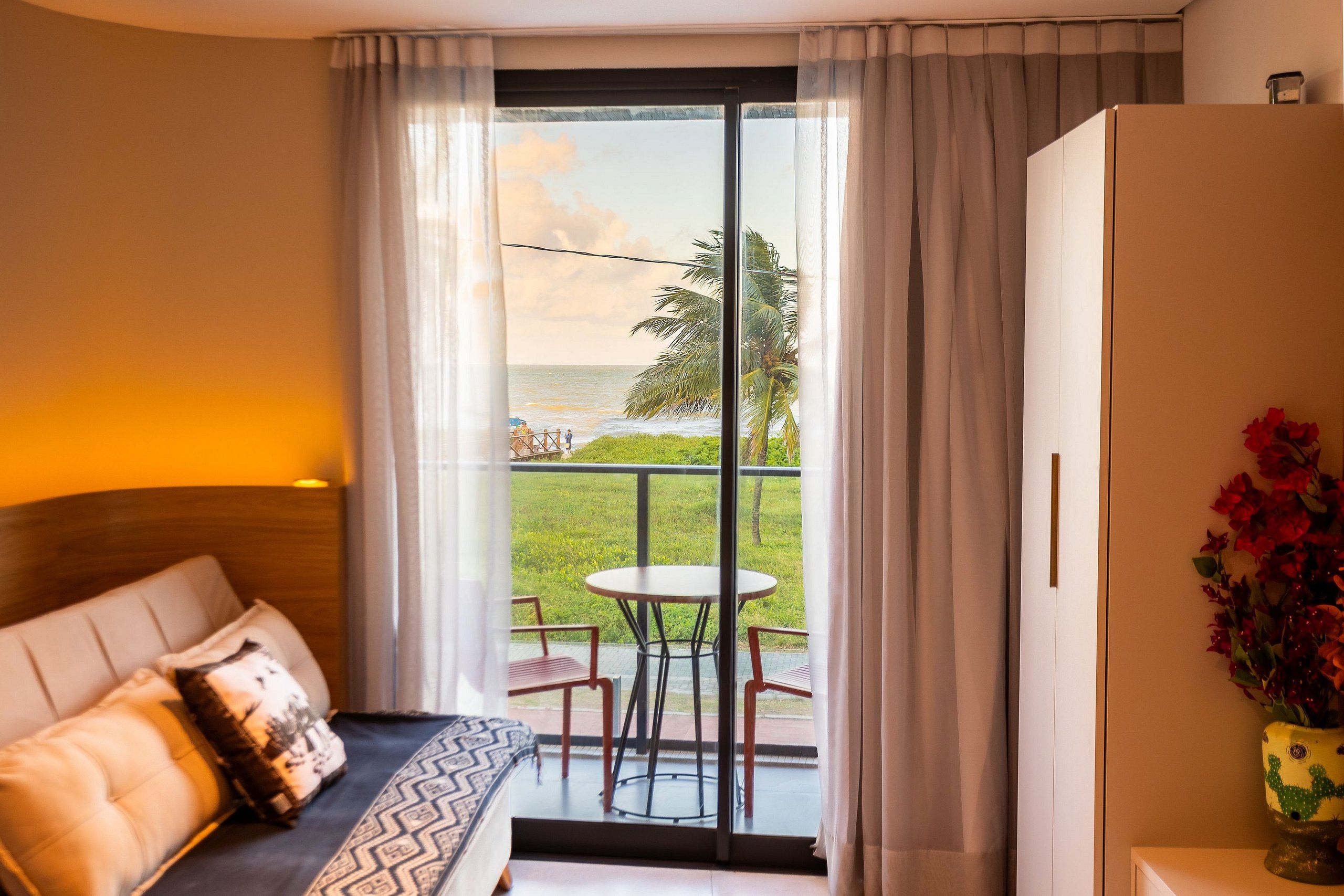 Quarto de hotel com cama, uma pequena mesa, cadeiras, varanda com vista para o mar e palmeiras, e um vaso em forma de cacto com flores.