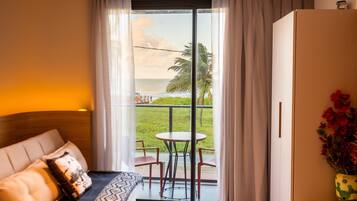 Quarto de hotel com cama, uma pequena mesa, cadeiras, varanda com vista para o mar e palmeiras, e um vaso em forma de cacto com flores.
