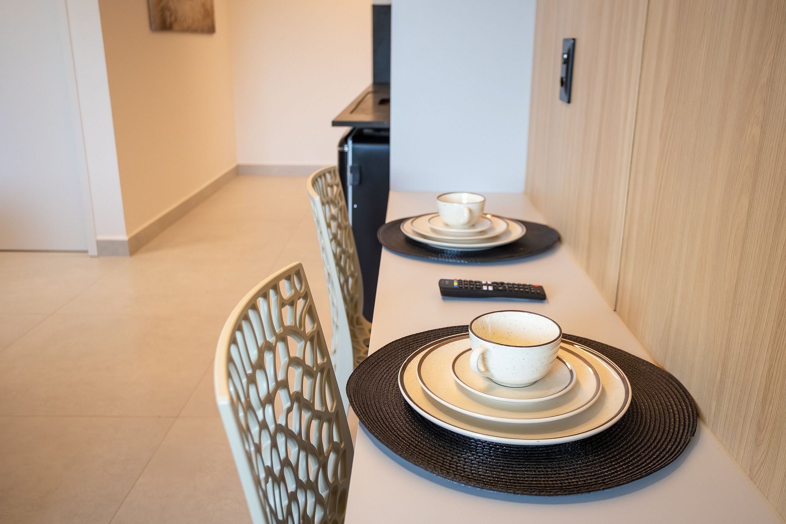 A dining table with a cup and saucer, a remote control, and a chair with a decorative backrest.