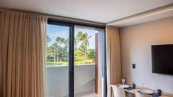 Habitación de hotel con balcón, zona de comedor con mesa para dos personas y televisor de pantalla plana montado en la pared.