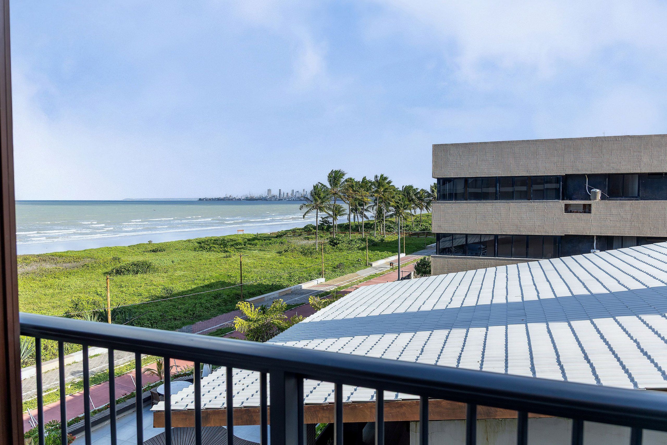 Vista de uma varanda sobre uma praia, área gramada e um edifício moderno com telhado plano.