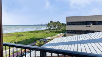 Vista de uma varanda sobre uma praia, área gramada e um edifício moderno com telhado plano.