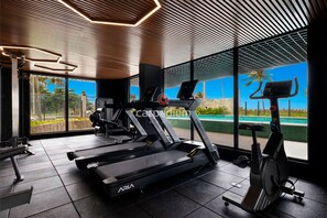 Un gimnasio moderno con cintas de correr, una elíptica y una máquina de remo, con ventanas amplias y vistas a una piscina.