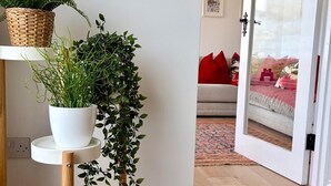 Een kamer met een houten vloer, een witte muur, een deur met ruit, een plank met planten en een rode kussen op een bank.