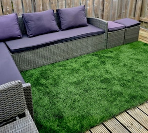 Un sofá modular con cojines, una zona de césped artificial verde y mobiliario de mimbre frente a un fondo de valla de madera.