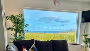 Een woonkamer met een groot raam, een grijze hoekbank en een potplant.