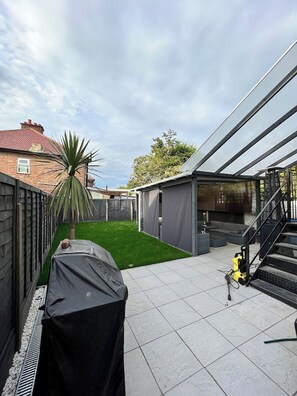 Un jardin avec un barbecue couvert, un palmier, un escalier et une terrasse couverte.