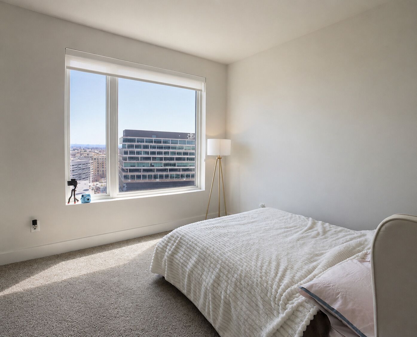 Un dormitorio con una ventana grande, una cama, una lámpara y vistas a un edificio.