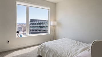 Un dormitorio con una ventana grande, una cama, una lámpara y vistas a un edificio.