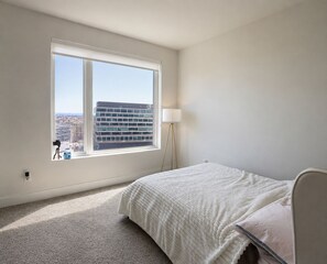 A bedroom with a large window, a bed, a lamp, and a view of a building.