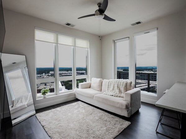A modern living room with a white sofa, large windows, a ceiling fan, and a view of the cityscape.