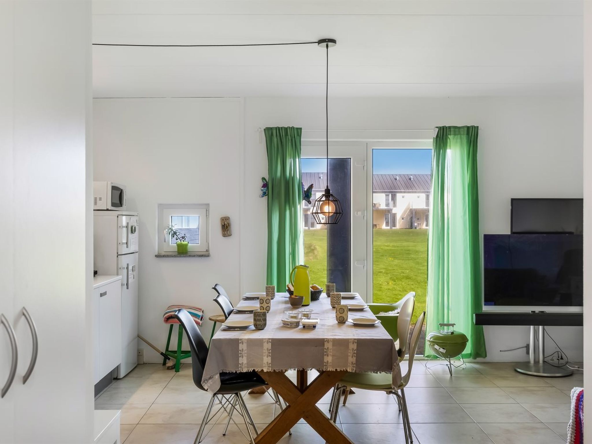 Un coin repas avec une table dressée pour quatre personnes, un téléviseur et une vue sur une cour verdoyante, visible à travers une porte.