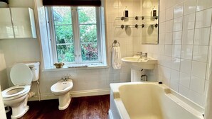 A bathroom with a bathtub, toilet, bidet, and sink, with a window providing a view of greenery outside. - Victorian Deco House - 5-bedroom house in Ventnor perfect for family getaways (Ventnor)