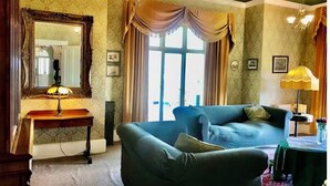 A room with a large mirror, a blue sofa, a table with a lamp, and a window with curtains. - Victorian Deco House - 5-bedroom house in Ventnor perfect for family getaways (Ventnor)