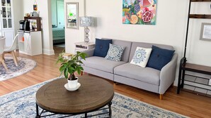 Una sala de estar con un sofá gris, cojines estampados, una mesa de centro redonda con una planta, una obra de arte colorida en la pared y una estantería.