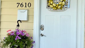 Una puerta blanca con el número amarillo '7610' encima, un buzón, una cesta de flores y un timbre.