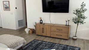 Ein Wohnzimmer mit einer Couch, einem Holzkabinett, einem an der Wand montierten Fernseher und einer Zimmerpflanze.