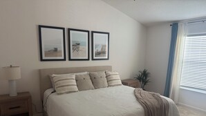 Ein Schlafzimmer mit einem Bett, Nachttischen, einer Lampe, einer Pflanze und drei gerahmten Bildern an der Wand.