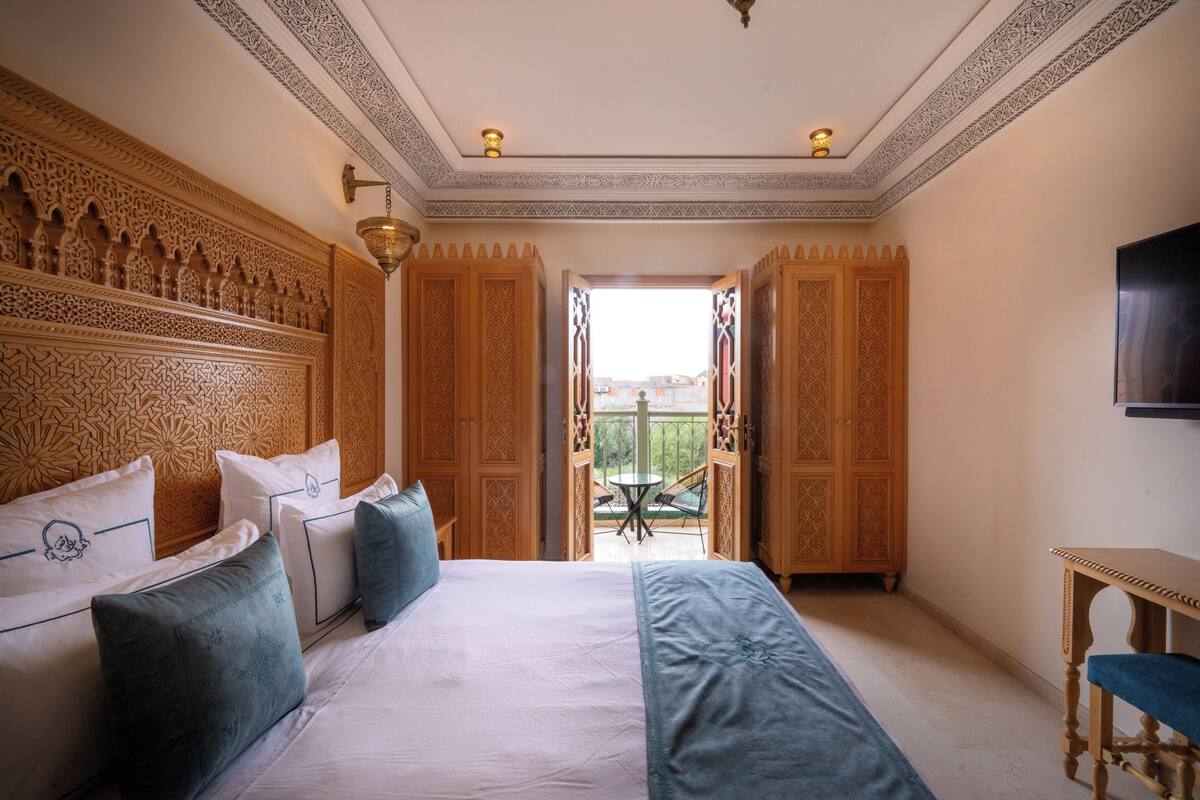 A bedroom with a large bed, ornate wooden headboard, a television, and a balcony with a table and chairs.