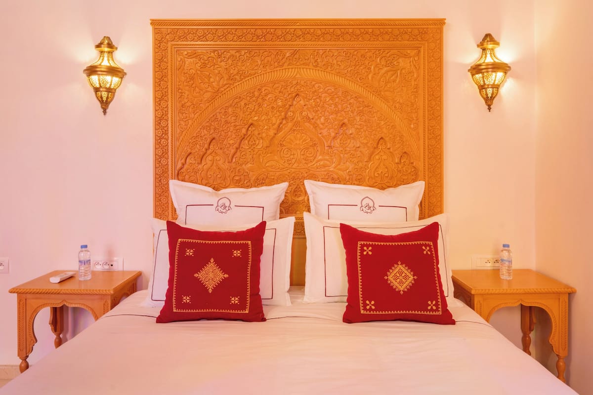 A hotel room with a large bed, ornate headboard, two bedside tables, and wall-mounted lamps.