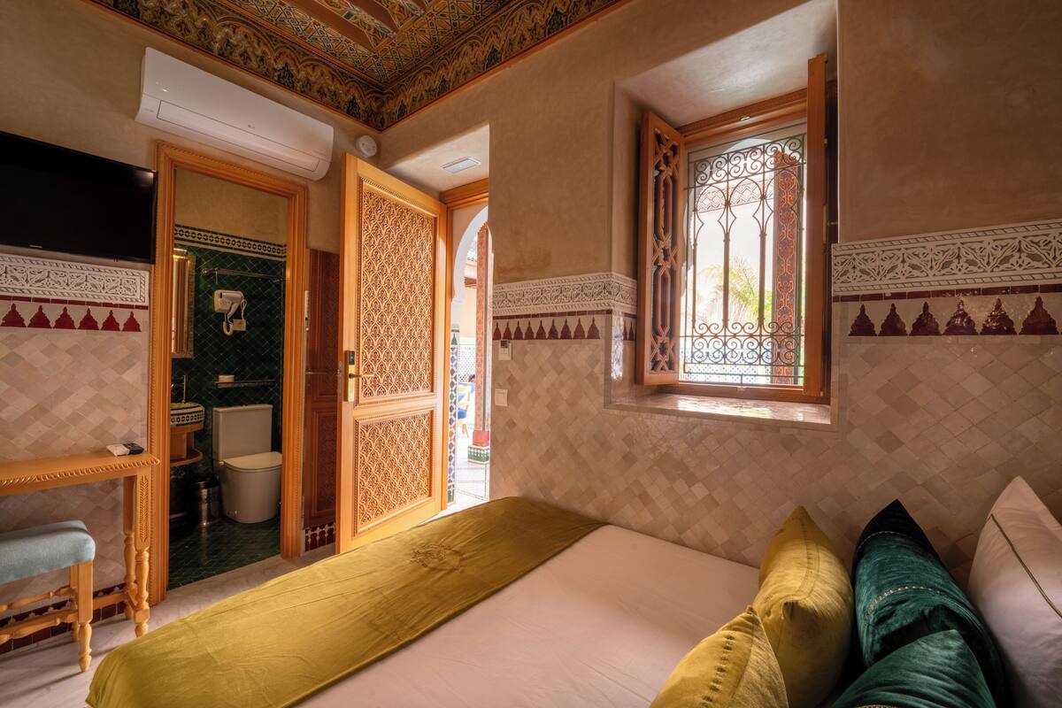 A room with a bed, a window with decorative ironwork, a door leading to a bathroom, and a small table with a chair.